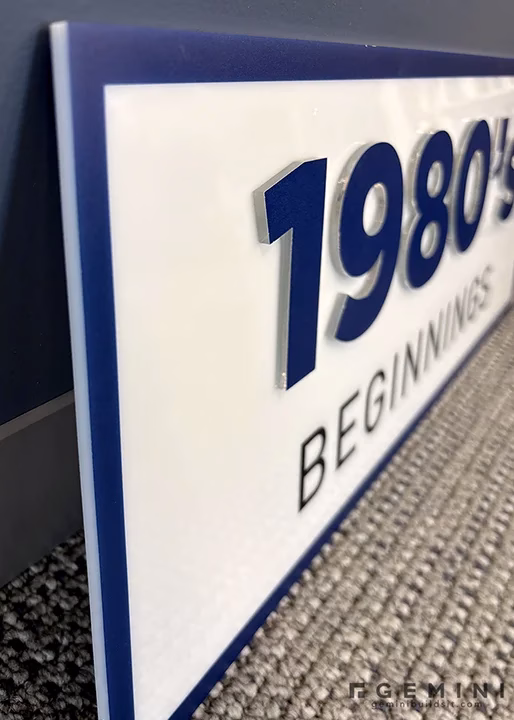 Close-up of a printed white acrylic decade marker reading 1980s Beginnings used on American Digital's corporate history wall
