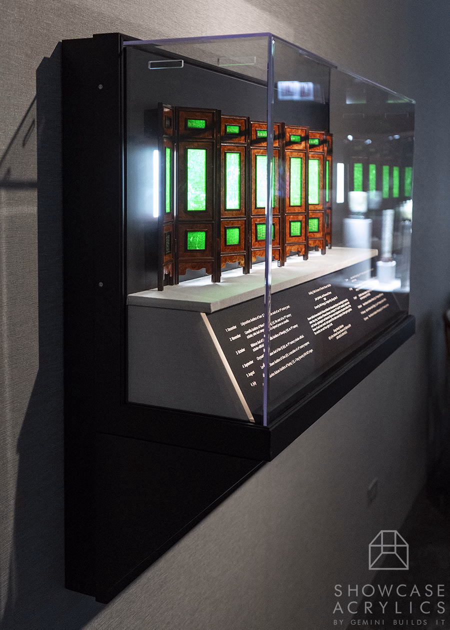 Display case housing a row of wooden frames with green illuminated panels in a museum exhibit, inside a glass enclosure.]','