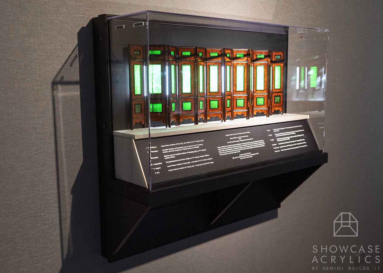 Display case mounted on a wall containing a row of ornate wooden panels with green glass inlays and an information plaque below.