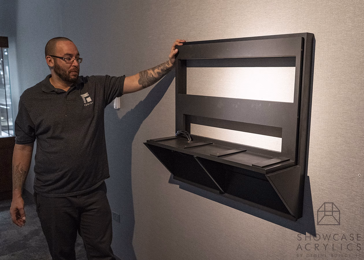Man with a beard and glasses stands beside a large black wall-mounted modern display unit with rectangular cutouts and an angled lower shelf.