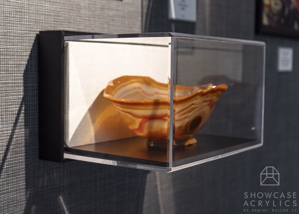 Clear wall-mounted acrylic display case containing a marbled orange and cream ceramic bowl, lit from the side.
